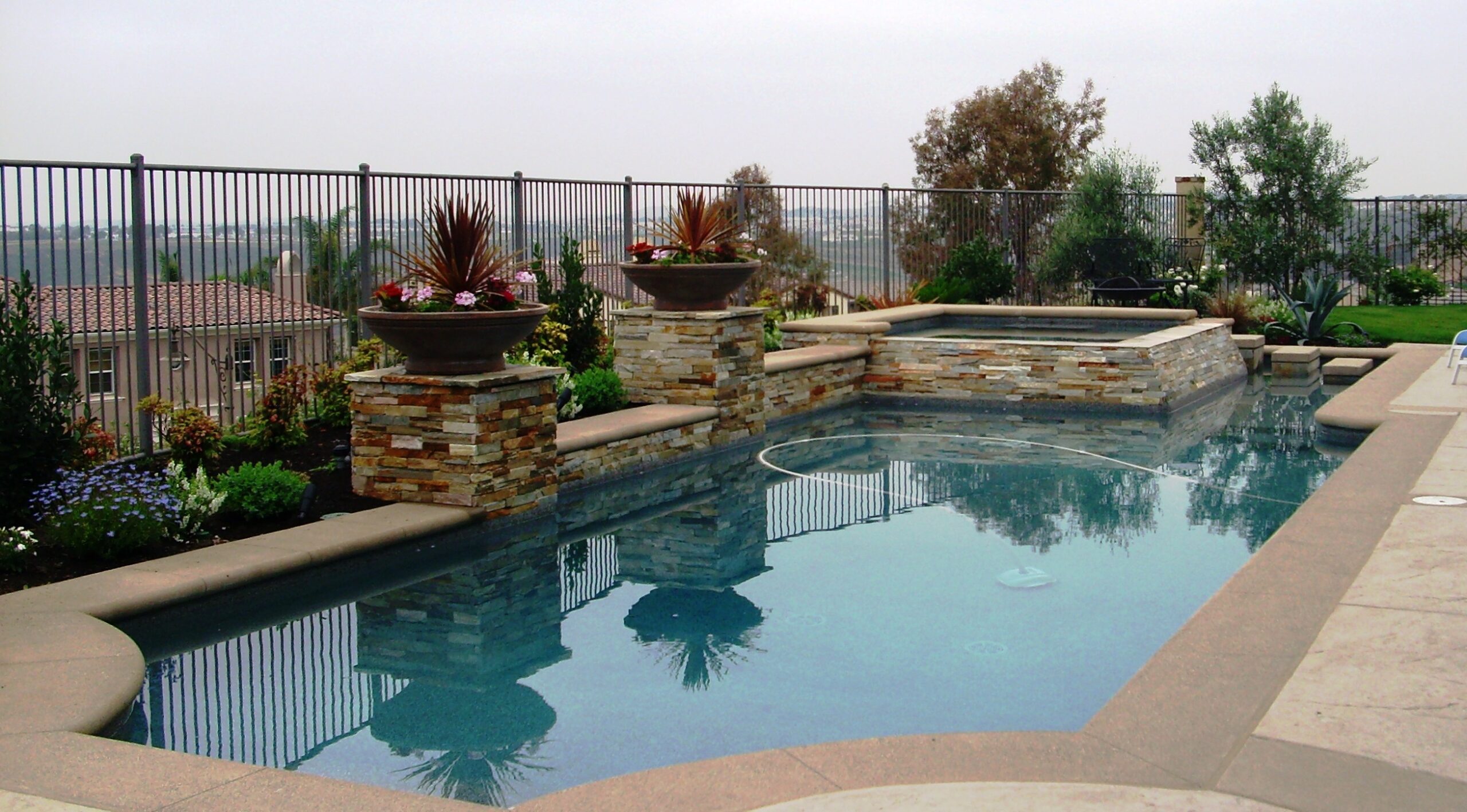 Kuyper Pool A modern outdoor pool with stone accents, surrounded by plants and flowers, set against a view of trees and distant buildings.